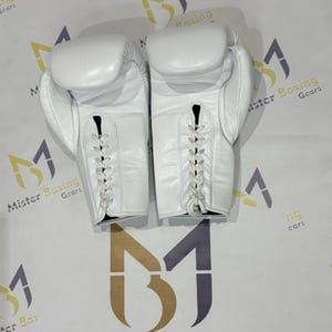 May include: A pair of white leather boxing gloves, laced up the front. The gloves are shown from a top-down perspective, with the laces visible. The gloves are on a white surface with the words "Mister Boxing Gears" printed on it.