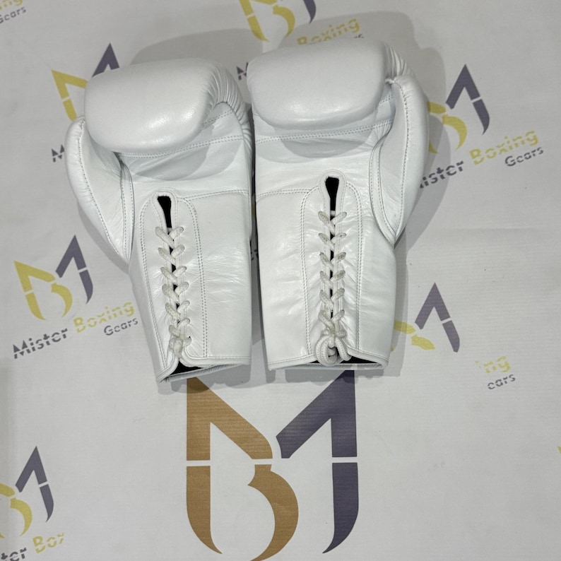May include: A pair of white leather boxing gloves, laced up the front. The gloves are shown from a top-down perspective, with the laces visible. The gloves are on a white surface with the words "Mister Boxing Gears" printed on it.