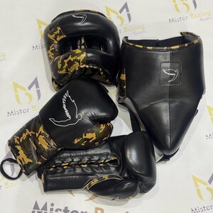 Puede incluir: Un conjunto de equipo de boxeo negro, que incluye un protector de cabeza, un protector facial y guantes. Los guantes y el casco presentan un patr&oacute;n de camuflaje en tonos marrones y verdes. Cada pieza tiene un logotipo de paloma blanca.