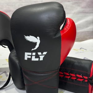May include: Black and red boxing gloves with the word "FLY" and a white graphic of a winged figure. The gloves have red stitching and are designed for boxing training and competition. The image also shows a head guard and a focus mitt.