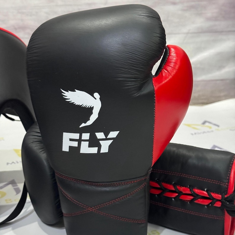 May include: Black and red boxing gloves with the word "FLY" and a white graphic of a winged figure. The gloves have red stitching and are designed for boxing training and competition. The image also shows a head guard and a focus mitt.