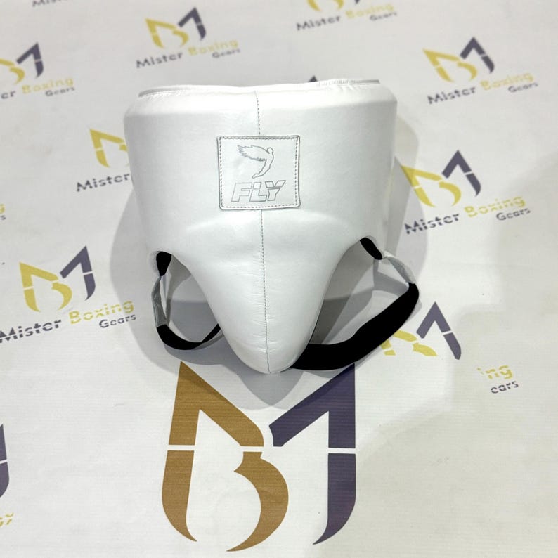 May include: White boxing groin protector with black straps. The protector has a stitched seam down the center and a small square patch with the word "FLY" and a bird logo. The product is designed for boxing and martial arts.