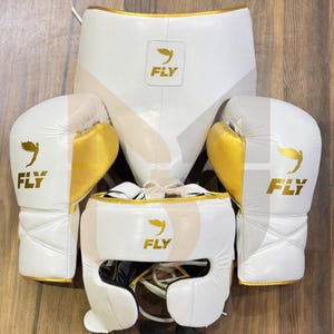 May include: A white boxing kit with gold detailing, comprising gloves, headgear, and a protective cup. Each item displays the word "FLY" and a bird logo. The set is designed for boxing training and competition.