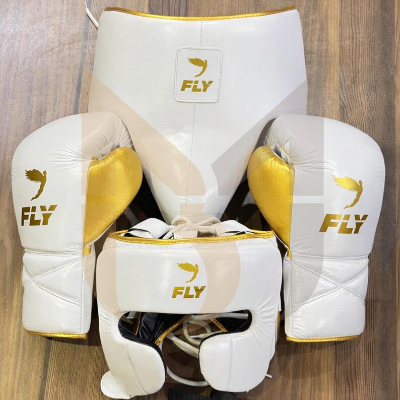 May include: A white boxing kit with gold detailing, comprising gloves, headgear, and a protective cup. Each item displays the word "FLY" and a bird logo. The set is designed for boxing training and competition.