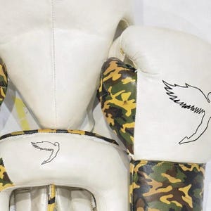 Puede incluir: Casco y guantes de boxeo blancos con detalles de camuflaje. Los guantes y el casco presentan un contorno negro de un p&aacute;jaro estilizado. Los guantes y el casco est&aacute;n hechos de un material blanco con detalles de camuflaje.