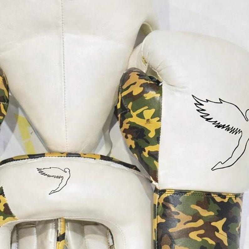 Puede incluir: Casco y guantes de boxeo blancos con detalles de camuflaje. Los guantes y el casco presentan un contorno negro de un p&aacute;jaro estilizado. Los guantes y el casco est&aacute;n hechos de un material blanco con detalles de camuflaje.