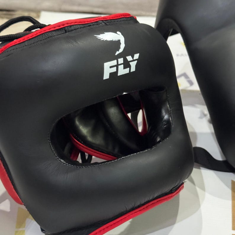 May include: Black boxing headgear with red trim and the word "FLY" in white. The headgear has a large opening for the face and is designed to protect the head during training or competition. A white bird graphic is above the word "FLY".