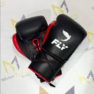 May include: A pair of black and red boxing gloves. The gloves have a white logo that says "FLY" with a bird graphic. The gloves are laced up with red laces and stitching. The gloves are made of leather.