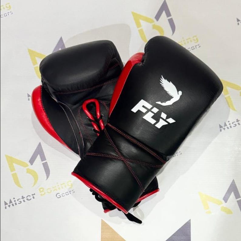 May include: A pair of black and red boxing gloves. The gloves have a white logo that says "FLY" with a bird graphic. The gloves are laced up with red laces and stitching. The gloves are made of leather.