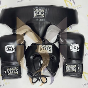 Peut inclure: Un ensemble d'équipement de boxe noir comprenant un protège-tête, des gants et une protection thoracique. Les gants et le casque affichent le logo "CLETO REYES" en blanc. L'ensemble est conçu pour l'entraînement et la compétition de boxe professionnelle.