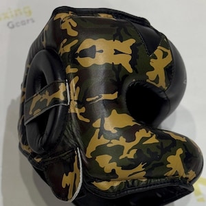 Puede incluir: Casco de boxeo de camuflaje con detalles en cuero negro. El casco presenta un patr&oacute;n de camuflaje verde, marr&oacute;n y beige. La protecci&oacute;n para los o&iacute;dos es negra y el casco tiene un cierre de correa. La imagen muestra el casco desde un &aacute;ngulo lateral.