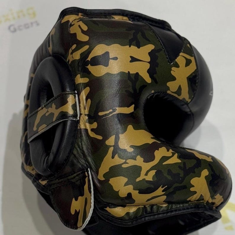 Puede incluir: Casco de boxeo de camuflaje con detalles en cuero negro. El casco presenta un patr&oacute;n de camuflaje verde, marr&oacute;n y beige. La protecci&oacute;n para los o&iacute;dos es negra y el casco tiene un cierre de correa. La imagen muestra el casco desde un &aacute;ngulo lateral.