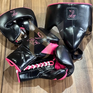 Könnte beinhalten: Ein Set schwarzer Boxausrüstung mit leuchtend pinken Akzenten und weißen Schnürsenkeln, bestehend aus einem Kopfschutz, einem Tiefschutz und einem Paar Boxhandschuhe. Jedes Teil zeigt das FLY-Logo mit einem Vogel-Symbol in Pink auf der Oberfläche.
