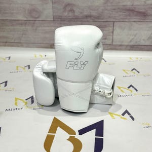 May include: A pair of white boxing gloves with the word "FLY" printed on the side. The gloves are made of a smooth material and have a stitched design. The background has a logo that says "Mister Boxing Gears".