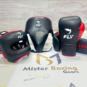 May include: A boxing gear set featuring a black headgear, a face protector, and boxing gloves with red accents. The gloves and headgear have the word "FLY" and a bird logo. The set is designed for protection during training and matches.