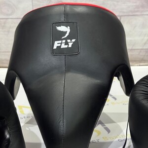 May include: Black boxing headgear with a red trim. The headgear has a square patch with the word "FLY" and a white bird logo. The headgear is designed to protect the head during boxing or other combat sports.