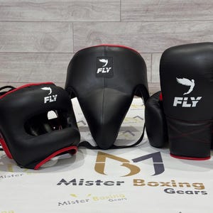 May include: A set of black boxing gear, including a head guard, face protector, and gloves. Each piece has red trim and the word "FLY" with a wing logo. The gear is displayed on a white surface with the text "Mister Boxing Gears".
