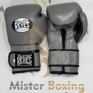 Peut inclure: Une paire de gants de boxe en cuir gris avec le logo Cleto Reyes sur la sangle du poignet et le dos de la main. La sangle du poignet comprend une petite étiquette blanche marquée 14 oz. Les gants présentent des bordures et des coutures noires.