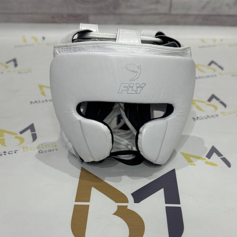 May include: A white boxing headgear with the word "FLY" printed on the front. The headgear has a padded design with openings for the eyes, ears, and mouth. The headgear is designed to protect the head during boxing or other combat sports.