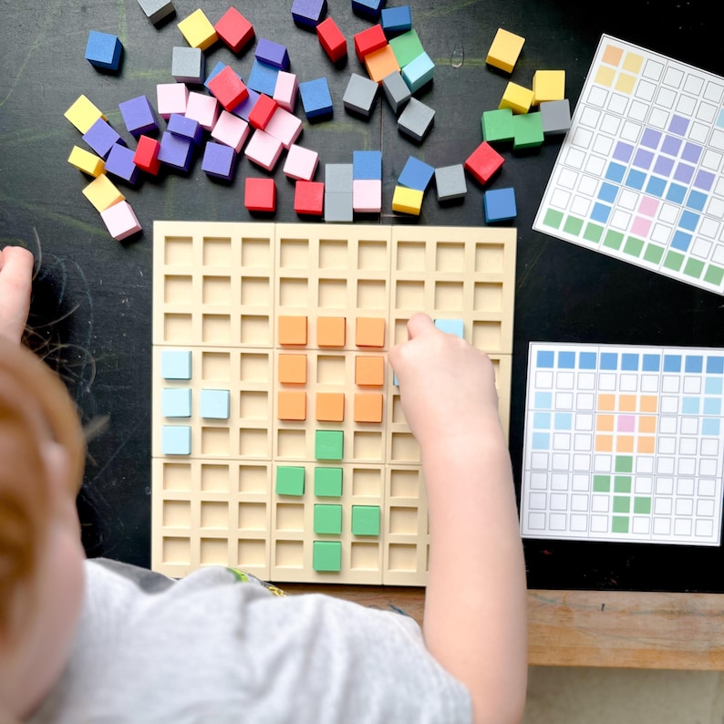 May include: A child's hand placing a blue square on a beige grid board. The board is part of a color matching game with colorful wooden blocks scattered around. Two printed pattern cards are also visible.