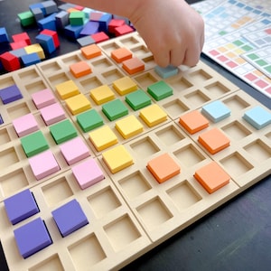 May include: A wooden educational toy with a grid of square cutouts, each holding a colorful square block. The blocks are in shades of purple, pink, green, yellow, orange, and blue. A hand is placing a blue block. Additional blocks are scattered nearby.