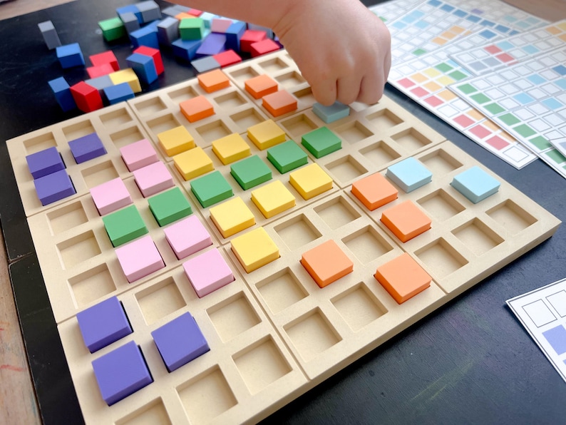 May include: A wooden educational toy with a grid of square cutouts, each holding a colorful square block. The blocks are in shades of purple, pink, green, yellow, orange, and blue. A hand is placing a blue block. Additional blocks are scattered nearby.