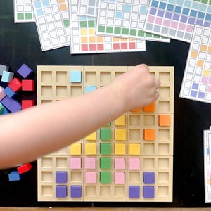 May include: A beige grid board with colorful square blocks being placed by a hand. Several printed sheets with colored squares are in the background, along with a pile of loose blocks. The blocks are various colors, including blue, red, orange, and yellow.