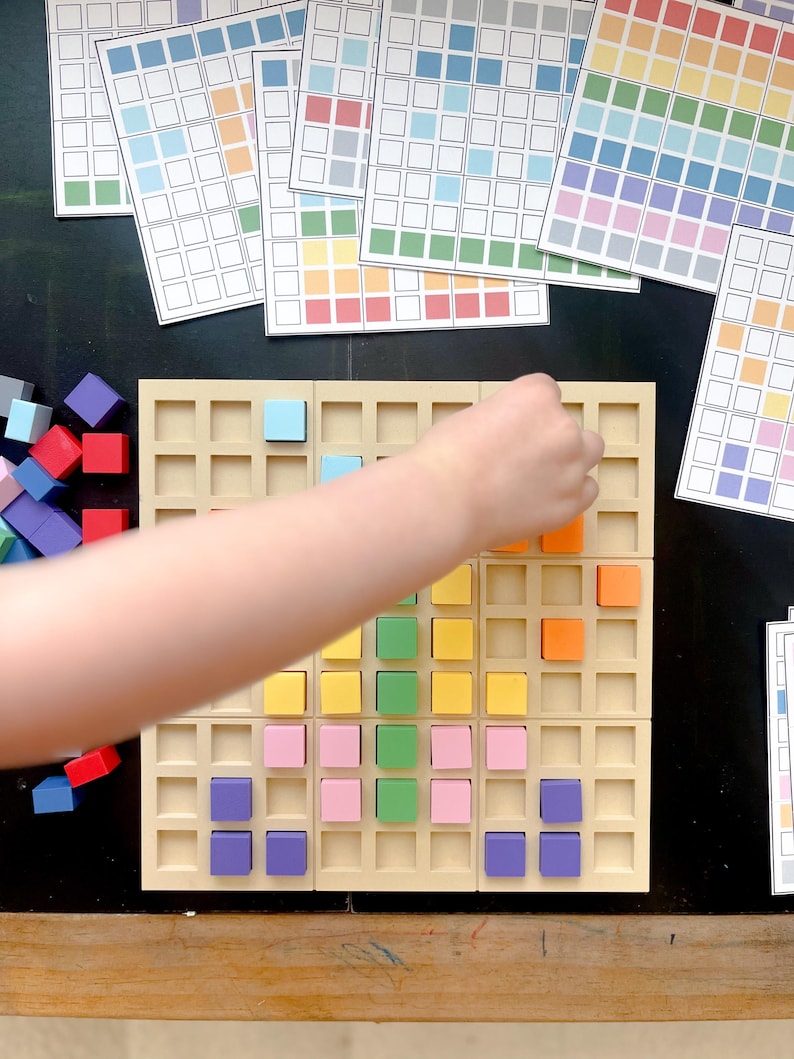 May include: A beige grid board with colorful square blocks being placed by a hand. Several printed sheets with colored squares are in the background, along with a pile of loose blocks. The blocks are various colors, including blue, red, orange, and yellow.