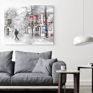 May include: A watercolour painting depicting a street scene with buildings and a pedestrian. The artwork is displayed on a white wall above a grey sofa. A modern white pendant light hangs from the ceiling. A small side table with a cup and saucer is in the foreground.
