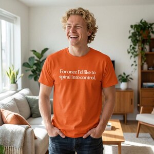Puede incluir: Camiseta naranja con el texto blanco "For once I'd like to spiral into control". La camiseta es de cuello redondo y está hecha de un material suave. La persona lleva vaqueros azules.