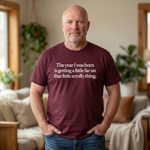 May include: A maroon t-shirt with the text "The year I was born is getting a little far on that little scrolly thing." The shirt is a crew neck and is being worn by a person.