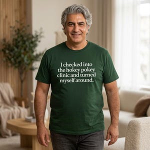 Op de afbeelding: Een donkergroen t-shirt met de tekst "I checked into the hokey pokey clinic and turned myself around" in het wit. Het shirt wordt gedragen door een persoon in een huiselijke omgeving.