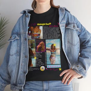 Op de afbeelding: Zwart T-shirt met de tekst "Midnight Sun" en albumafbeeldingen. Het shirt toont een lijst met songtitels en een foto van een vrouw in badpak. Het T-shirt wordt gedragen met een blauw spijkerjasje.