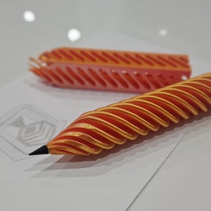 May include: An orange and gold pencil with a unique spiral design, resting on a white surface. A matching orange pencil holder is in the background. A sketch is visible on the paper.