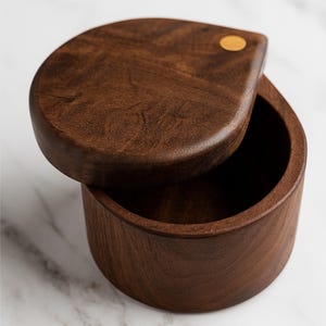 May include: A dark brown wooden salt cellar with a teardrop-shaped lid. The lid is slightly ajar, revealing the interior of the round container. A small, round, gold-coloured accent is on the lid. The wood grain is visible.