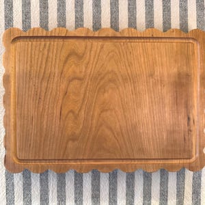 May include: A rectangular wooden cutting board with a scalloped edge. The board is made of light brown wood and has a recessed area. The cutting board is on a striped fabric background.