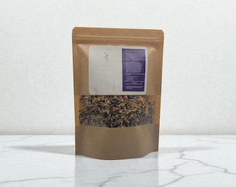 May include: A brown paper bag with a clear window displaying dried herbs. The bag has two labels, one with the text "LA TEE" and a small illustration. The other label is purple with text. The bag is sitting on a white marble surface.