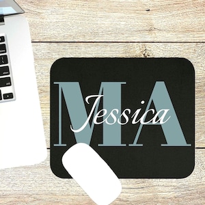 May include: Black mousepad with the letters "MA" in a light blue color and the name "Jessica" in white script. A white computer mouse is next to the mousepad. The background is a light brown wood grain.