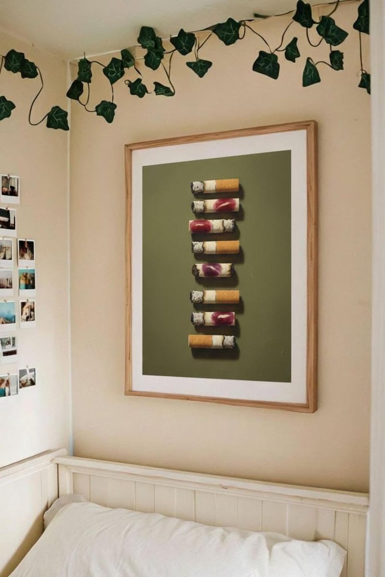The framed lipstick cigarettes print hanging on a beige wall above a bed decorated with artificial ivy.