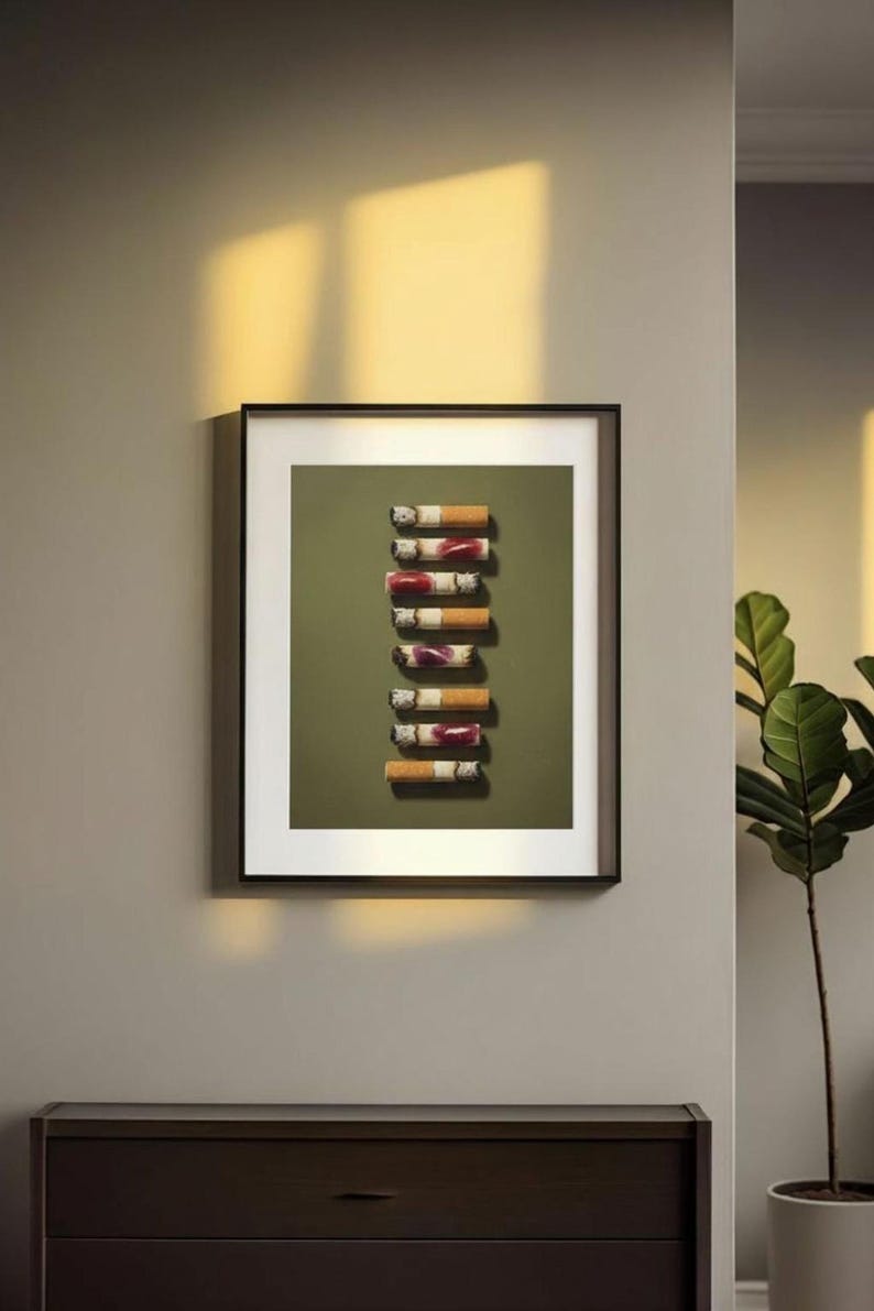 A black-framed print of the lipstick cigarettes artwork hanging on a grey wall with warm sunlight washing over it.