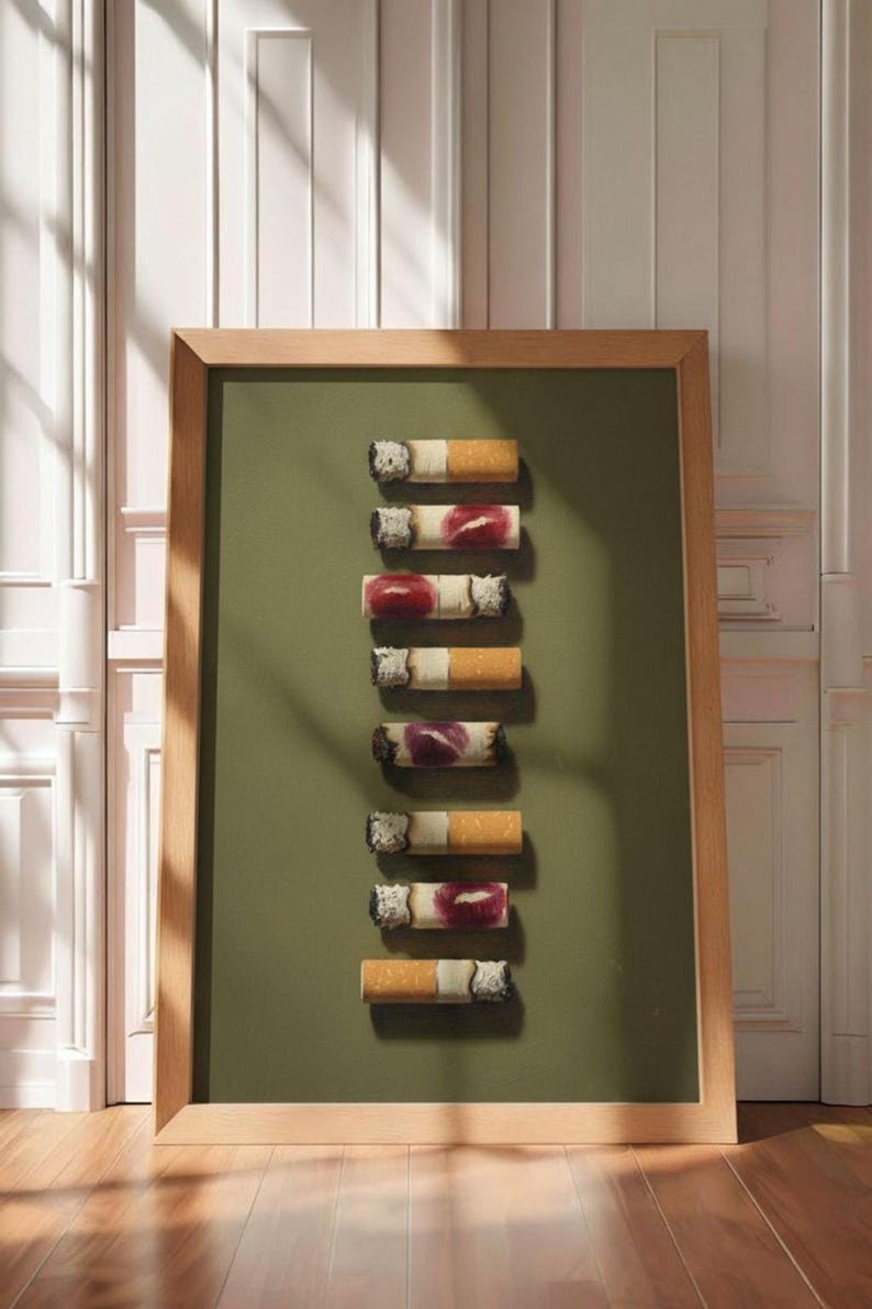 A large framed print of the lipstick cigarettes artwork leaning against a white paneled wall on a wood floor.
