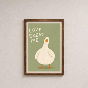 May include: A framed art print featuring a cartoon goose with orange feet and beak, set against a muted green background. The text "LOVE BREAD ME" is written in white above the goose. The frame is a dark brown color.