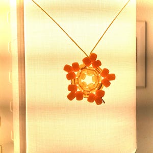 May include: A handmade necklace featuring a star-shaped pendant. The pendant is crafted from orange and yellow beads, strung on a thin, yellow cord. The necklace is displayed against a soft, illuminated background.
