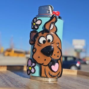 Könnte beinhalten: Ein dekoratives Feuerzeug mit einem Cartoon-Hund-Design. Das Feuerzeug hat einen hellblauen Hintergrund mit einem braunen Hunde-Design, einer rosa herzförmigen Zunge und Blumenakzenten. Das Feuerzeug steht auf einer Holzoberfläche vor blauem Himmel.