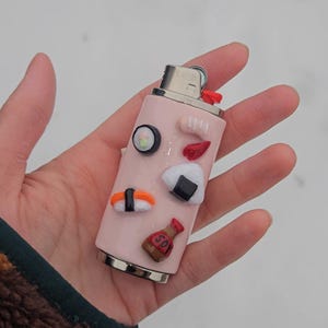 May include: A pink lighter decorated with sushi-themed embellishments. The lighter features various sushi pieces, including a roll, nigiri, and onigiri, along with a small soy sauce bottle. The lighter is held in a hand against a snowy background.