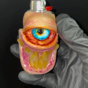 May include: A custom lighter case featuring a sculpted, fleshy creature design with a prominent, detailed blue eye and a wide, open mouth revealing yellowed teeth. The surface has pink and peach tones, with a metallic silver lighter mechanism on top.