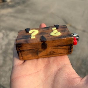 May include: A rectangular lighter case with a brown wood-grain finish, featuring two raised yellow question marks on the top surface and a dark brown latch detail. The metal ignition mechanism and a small red button are visible on the right side.