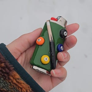 May include: A green lighter decorated with billiard ball designs. The lighter features a pool cue and several billiard balls, including the 5-ball, 8-ball, and 1-ball. The lighter is held against a snowy background.