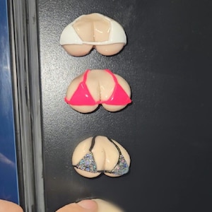 May include: Three novelty magnets shaped like a pair of breasts, each wearing a different bikini top. The tops are white, hot pink, and silver glitter. The magnets are displayed on a black surface.