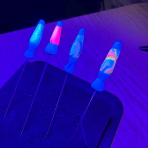 Op de afbeelding: Vier miniatuur bedels in de vorm van lavalampen met blauw oplichtende voetjes en stelen. Elke bedel heeft een uniek intern kleurpatroon, waaronder effen roze, wit en veelkleurige wervelingen, allemaal verlicht onder ultraviolet licht.